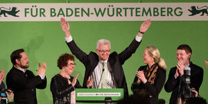 Winfried Kretschmann hebt umringt von applaudierenden Parteikolleg_innen die Arme.