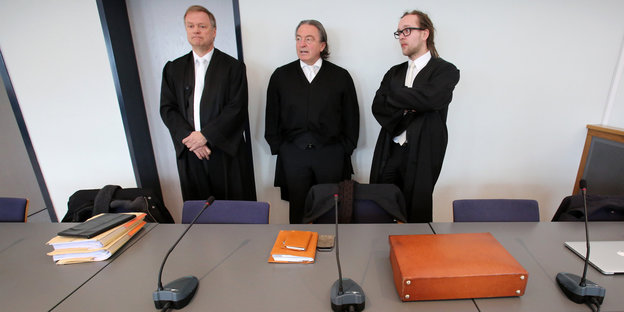 Drei Juristen in schwarzen Roben lehnen hinter einem leeren Tisch an der Wand.