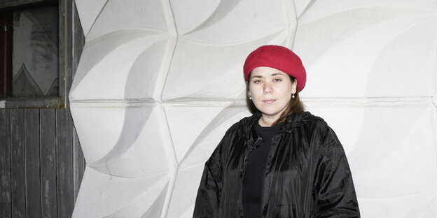 Stefanie Sargnagel in schwarzer Jacke und roter Mütze lehnt an einer grau-weißen Wand.