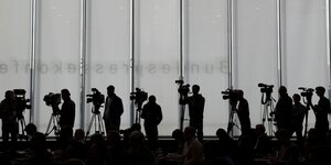 Journalisten mit Kameras stehen vor einer grau-weiße Wand.