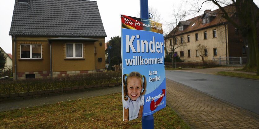 Ein AfD-Plakat, das Kinder willkommen heißt, hängt an einem blauen Pfahl, dahinter graubraune Häuser.