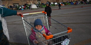 Ein Kind sitzt in enem Einkaufswagen, dahinter eine Reihe Zelte.