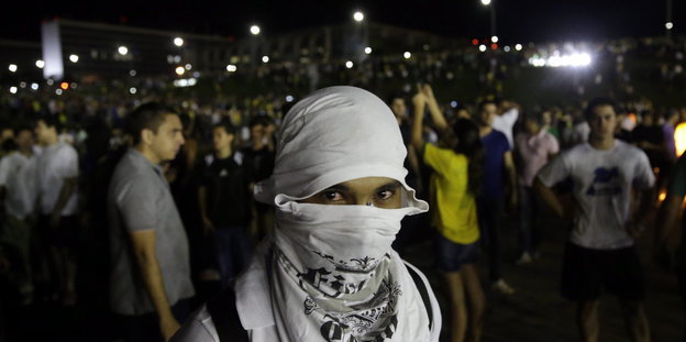 Ein Mensch hat sein Gesicht mit einer weißen Kopfbedeckung und einem weißen Tuch maskiert. Dahinter stehen Demonstrant_innen.