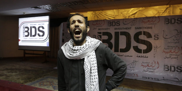 Ein Mann in dunkler Kleidung und Kufiya steht schreiend vor dem Schriftzug „BDS“ an einer Wand.