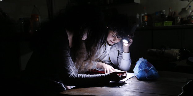 zwei Menschen sitzen mit Taschenlampe am Tisch und lesen etwas, sonst ist es dunkel