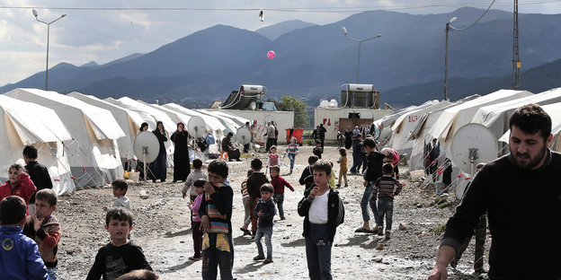 Kinder und ein Mann sind in einem Flüchtlingslager zu sehen, im Hintergrund Berge