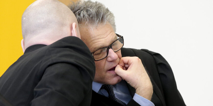 Ein Mann mit Glatze beugt sich zu einem anderen Mann mit grauen Haaren und Brille