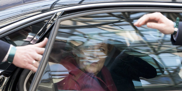 Angela Merkel sitzt im Auto. Eine Hand versucht ihr die Tür aufzuhalten.