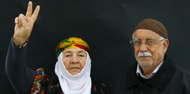 Eine Frau mit Kopftuch zeigt ein Peace-Zeichen mit zwei Fingern und ein Mann steht neben ihr.
