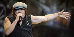 ein Mann in schwarzem Jeanshemd mit abgeschnittenen Ärmeln, Schiebermütze, in der einen Hand ein Mikrofon, den anderen Arm ausgesteckt