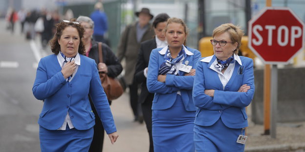 Drei Flugbegleiterinnen in blauen Kostümen, zwei davon mit verschränkten Armen, verlassen den Flughafen in Brüssel
