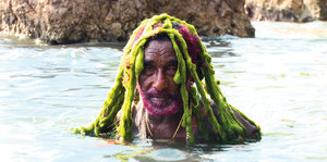 Ein Mann im Wasser, er hat einen pinken Bart und grüne Rastazöpfe