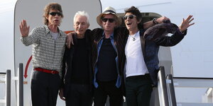 Die vier Mitglieder der Rolling Stones stehen oben auf einer Gangway vor der Eingangstür zum Flugzeug und winken