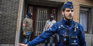 Zwei vermummte Männer stehen in einer Tür, vor ihnen ein Polizeibeamter in blauer Uniform