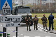 Schwer bewaffnete Soldaten riegeln eine Straße ab. Im Vordergrund: Hinweisschild zum Flughafen. Im Hintergrund: Militärlaster