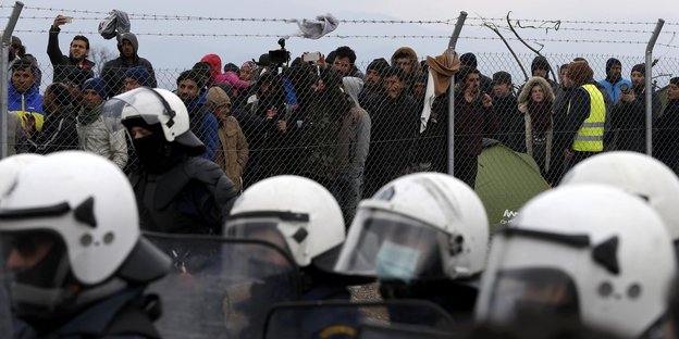 Menschen hinter einem Zaun, im Vordergrund Polizisten mit weißen Helmen