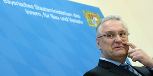 Ein Mann mit Brille und grauen Haaren, Bayerns Inneminister Herrmann, steht vor einer blauen Tafel