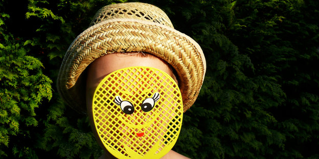 Eine Person mit Hut hält sich eine gelbe Fliegenklatsche vor ihr Gesicht, auf der Klatsche ist ein Smiley