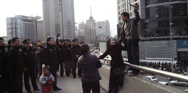 Ein Mann steht auf einem Brückengeländer vor Polizisten. Er hält eine Frau fest und in der anderen Hand ein Beil.
