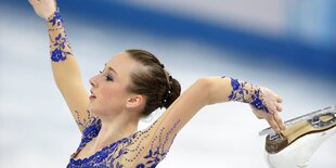 Die Einkunstläuferin hält mit einer Hand die Schiene ihres Eislaufschuhs fest, die andere Hand hält sie in die Höhe. Sie trägt ein blaues Kostüm und ist geschminkt