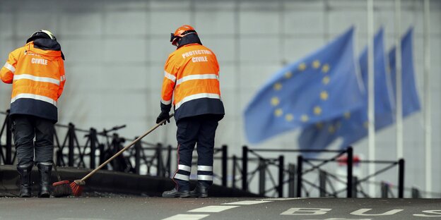Zwei Menschen mit Warnwesten fegen die Straße vor EU-Flaggen