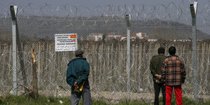 Drei Männer stehen vor der mit Stacheldraht gesicherten Grenze zwischen Griechenland und Mazedonien.