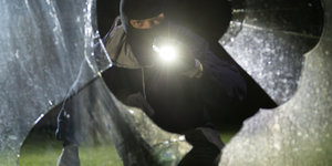 Ein maskierter Mann leuchtet mit Taschenlampe in ein zerbrochenes Fenster