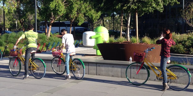 Drei Menschen auf knallbuten Fahrrädern, im Hintergrund eine grüne Android-Plastikfigur Drei Menschen auf knallbuten Fahrrädern, im Hintergrund eine grüne Android-Plastikfigur