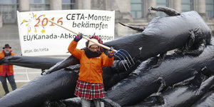 Frau sticht mit Mistgabel in schwarze Gummiwülste, die ein Monster darstellen, dahinter ein Transparent mit der Aufschrift "Ceta-Monster bekämpfen"