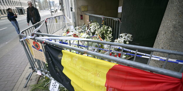 Belgische Flagge hängt an einem Gitter, dahinter Blumen, ein Mann guckt auf die Szenerie