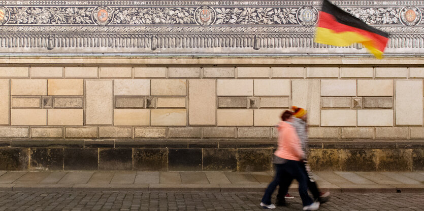 Zwei Menschen, die nicht im Detail zu erkennen sind, laufen eine Straße entlang. Sie tragen eine Deutschlandfahne