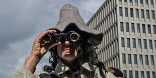 Ein Mann mit Schlapphut und Fernglas vor einem Hochhaus