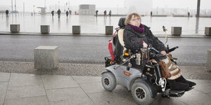 Eine Frau im Rollstuhl, dahinter das verregnete Berlin