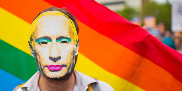 Mann mit geschminkter Putinmaske vor einer Regenbogenfahne