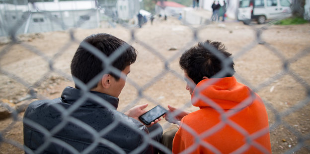 zwei Männer hinter einem Zaun schauen auf ein Smartphone