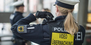 Eine Polizistin hält ihre Hand an eine Bodycam auf ihrer Schulter. Auf ihrem Rücken steht „Videoüberwachung“