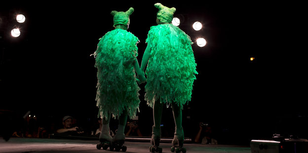 Zwei Personen in grünen Kostümen auf Rollerblades stehen auf einer Bühne und halten Händchen. Man sieht sie von hinten