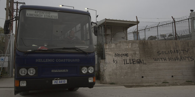 Ein Bus der griechischen Küstenwache verlässt das Lager. Graue Mauern, an denen „No one is illegal“ und „Solidarity with Migrants“ steht, darüber erhebt sich Stacheldraht