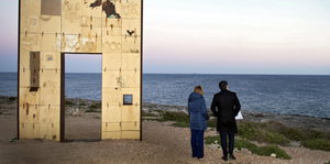 Zwei Menschen stehen neben einem freistehenden Steintor, dahinter das Meer