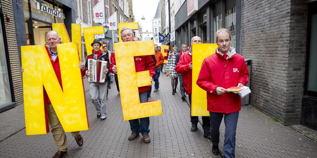 Mehrere Männer in roten Anoraks tragen Buchstaben, die das Wort NEE formen, durch eine Straße