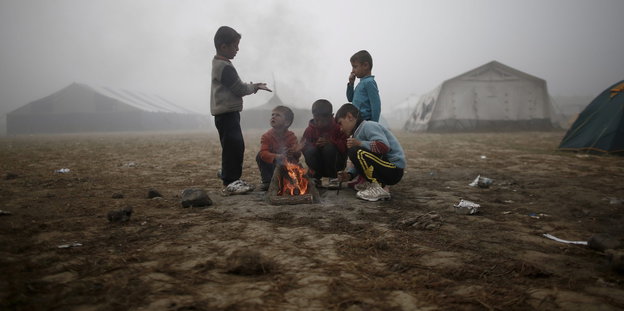 Kinder um ein kleines Lagerfeuer vor nebliger Zeltstadt