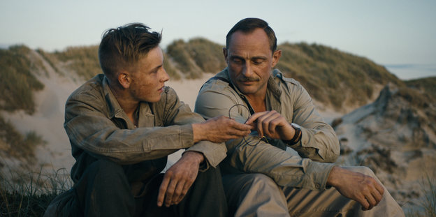 Zwei Männer sitzen an einer Düne und reichen sich einen Draht