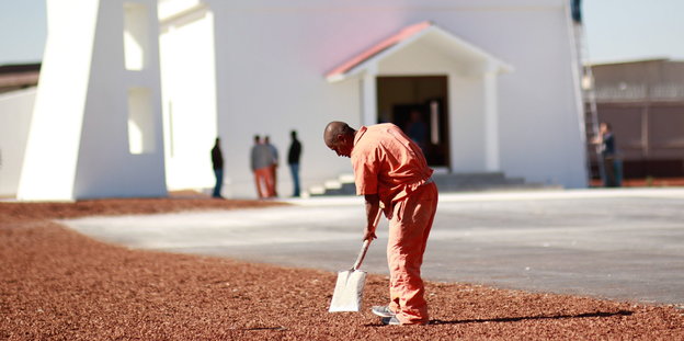 Ein Mann in oranger Häftlingskleidung mit einer Schaufel, im Hintergrund ein weißer Kirchenbau