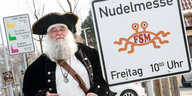 ein Mann mit weißem Bart und schwarzem Hut neben einem Schild, auf dem „Nudelmesse, Freitag, 10:00 Uhr“ und das Symbol eines Spaghettimonsters zu sehen ist