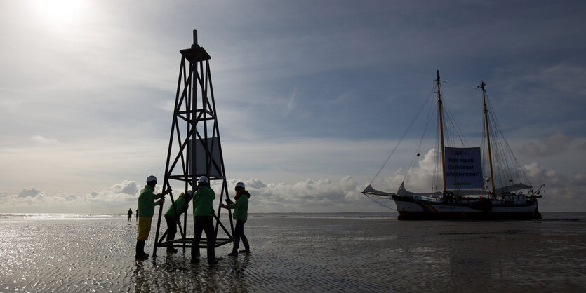 Mehrere Personen stehen um einen Holzbohrturm herum, im Hintergrund liegt ein Schiff im Meer