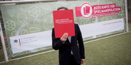 Ein Mann steht vor einem Fußballtor, in dem ein Banner mit der Aufschrift "Rote Karte für Spielmanipulation" hängt und hält eine rote Karte, die sein Gesicht verdeckt