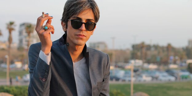 Payam Feili sitzt mit Sonnenbrille, Zigarette in der Hand, blau lakierten Fingernägeln und einem blauen Ring vor einer Stadtlandschaft. Am Hals ein Davidstern-Tattoo
