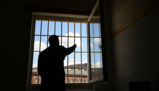 Ein Mensch steht an einem vergitterten Fenster, dahinter ist blauer Himmel zu sehen