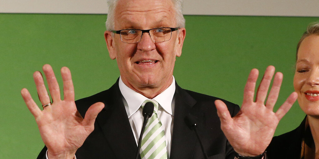 Ein Mann, es ist Winfried Kretschmann, hebt die Hände in die Luft und steht vor einem grünen Hintergrund