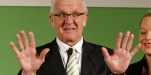 Ein Mann, es ist Winfried Kretschmann, hebt die Hände in die Luft und steht vor einem grünen Hintergrund